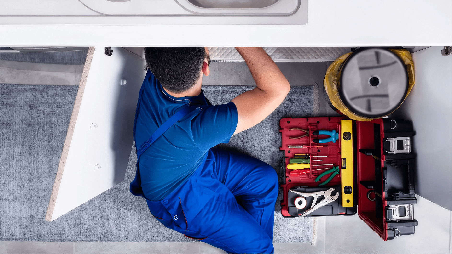 Plumber working beneath a kitchen sink with an open toolbox and plumbing tools, performing under-sink plumbing repairs or maintenance in a residential home.