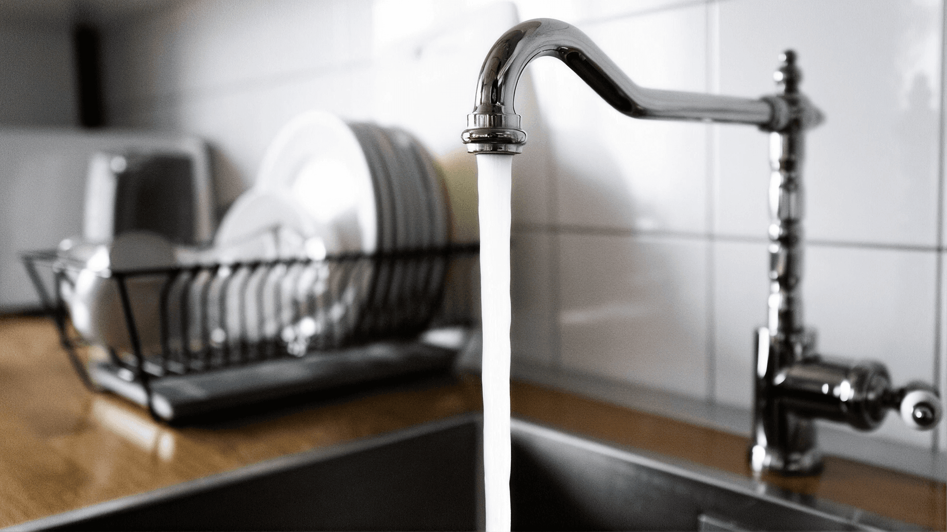 Clean kitchen faucet dispensing a steady stream of water into a stainless steel sink, with a dish drying rack holding plates and bowls on a wooden countertop in the background.