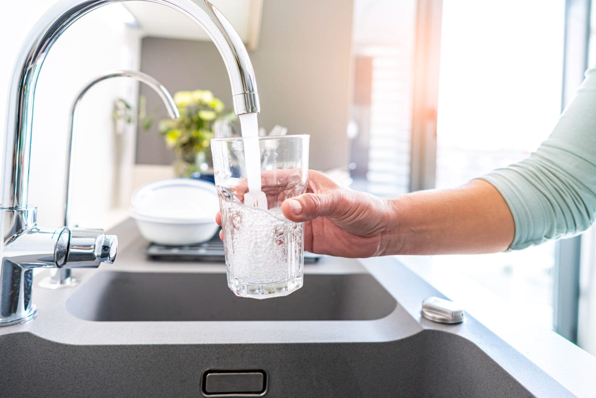 Hand filling a clear drinking glass with fresh tap water from a modern kitchen faucet over a stainless steel sink in a bright, sunlit kitchen.